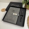 Set libreta con broche magnético plateado y bolígrafo negro en caja, mostrado junto a una planta y un objeto de madera sobre un escritorio.