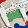 Un calibrador mide una "Placa de identificación para mascota personalizada - Hueso - Aluminio" verde, rodeada de coloridas placas de identificación sobre una superficie beige.