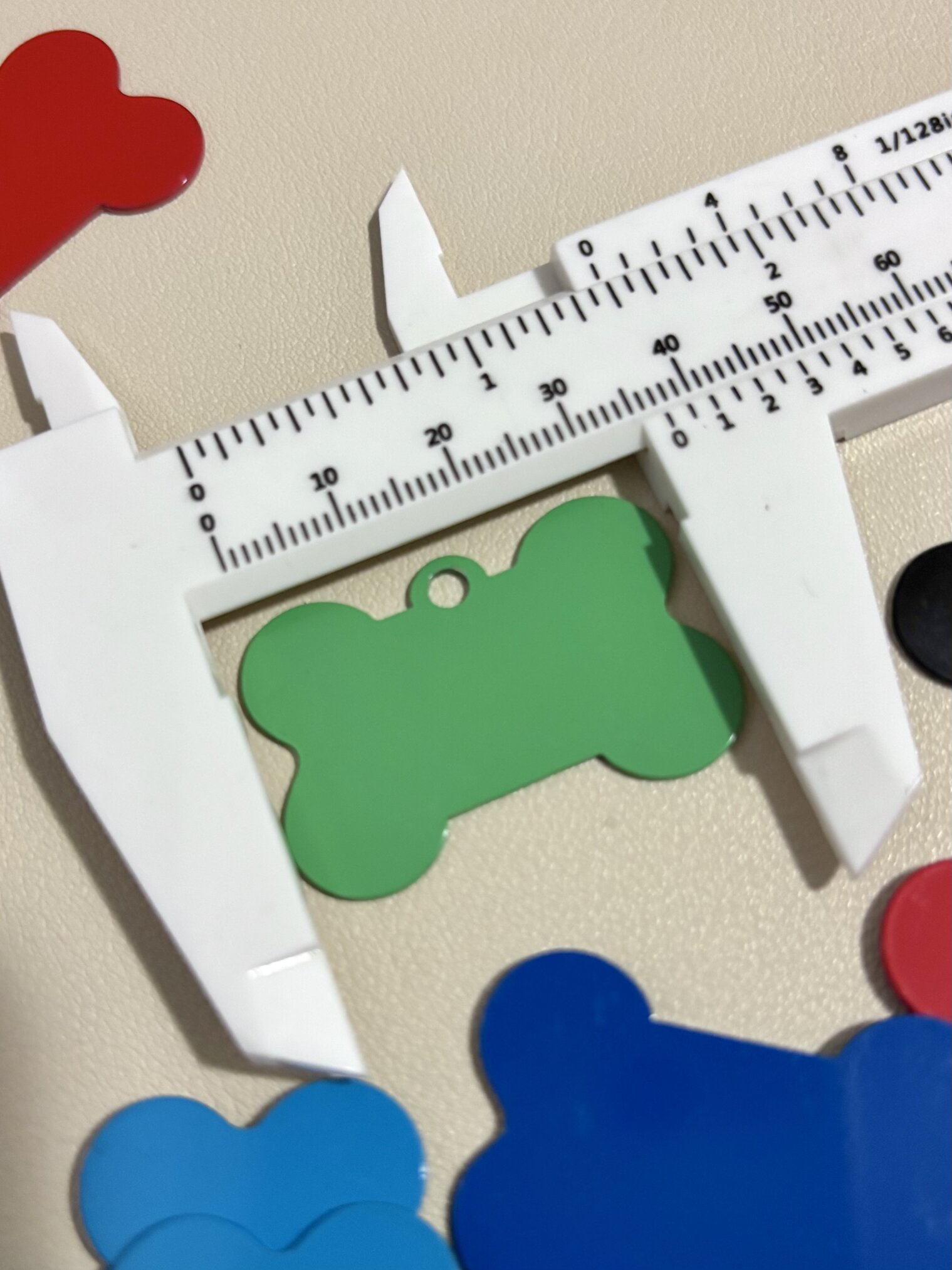 Un calibrador mide una "Placa de identificación para mascota personalizada - Hueso - Aluminio" verde, rodeada de coloridas placas de identificación sobre una superficie beige.