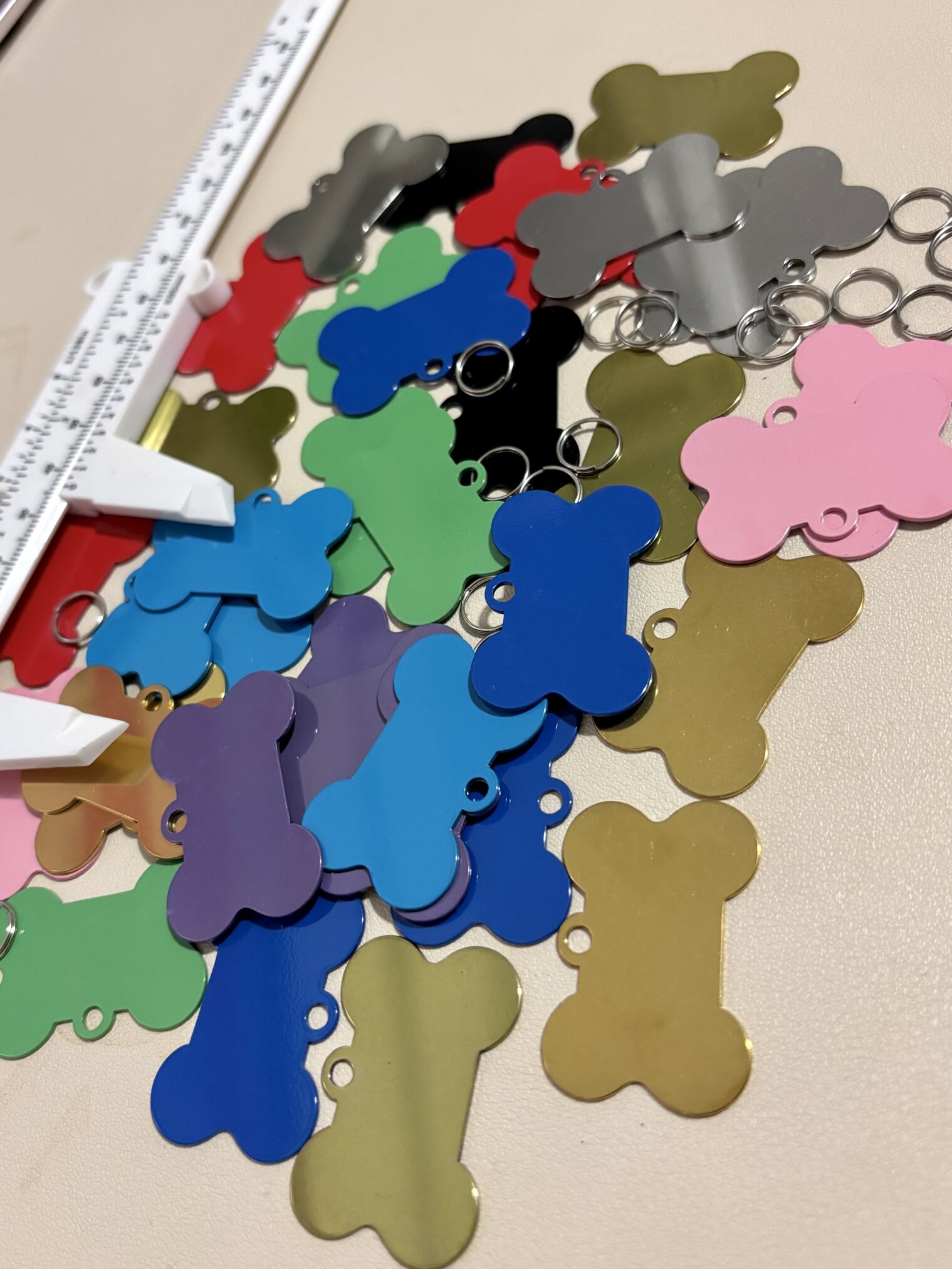 Placas de identificación para mascota personalizadas en forma de hueso de aluminio, algunas con llavero, están esparcidas junto a un calibrador blanco sobre una superficie beige.
