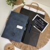 Set personalizado con bolígrafo y libreta con broche plateado, acompañado de una tarjeta de agradecimiento, una bolsa de papel y una caja redonda de madera, todo ello presentado sobre un mantel individual de tela: un set pensado para cualquier ocasión.