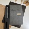 Un conjunto personalizado muy bien presentado, compuesto por un bolígrafo y un cuaderno con broche plateado, que incluye un cuaderno gris con nombre grabado en la portada, un bolígrafo personalizado y una bolsa de regalo oscura, todo ello expuesto sobre un escritorio.