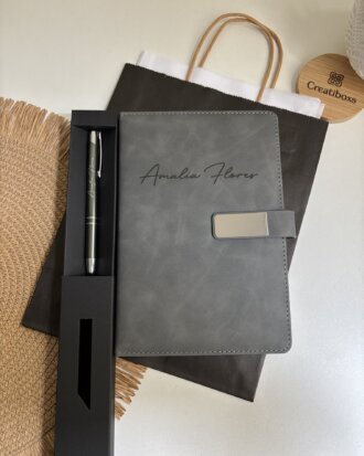 Un conjunto personalizado muy bien presentado, compuesto por un bolígrafo y un cuaderno con broche plateado, que incluye un cuaderno gris con nombre grabado en la portada, un bolígrafo personalizado y una bolsa de regalo oscura, todo ello expuesto sobre un escritorio.