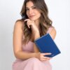 Una mujer con un vestido rosa sostiene la «Agenda diaria 2027» con bolígrafo, estuche y cuaderno, y sonríe pensativa a la cámara, como si estuviera a punto de anotar sus planes.