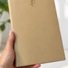 Una mano sostiene una agenda personalizada diaria 2027 profesional de color beige con números dorados en la portada; al fondo se ve una planta difuminada.