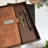 Conjunto compuesto por una libreta con cierre magnético plateado y un bolígrafo, presentado en una caja cuadrada; incluye un posavasos y una planta.