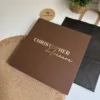 Conjunto compuesto por una libreta con cierre magnético plateado y un bolígrafo, presentado en una caja cuadrada sobre un mantelito, junto a una planta y un posavasos.