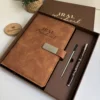 Conjunto compuesto por una libreta con cierre magnético plateado y un bolígrafo, presentado en una caja cuadrada, con una planta en maceta al fondo.