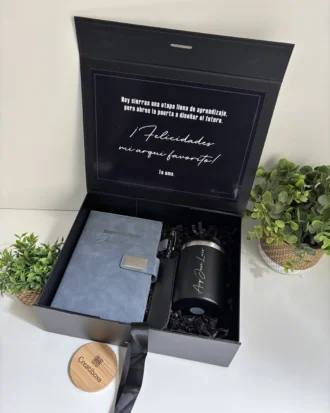 Kit personalizado en caja negra con taza termo negra, libreta azul con broche metálico y bolígrafo.
